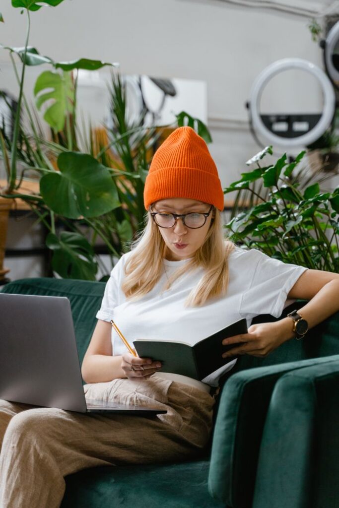 A woman wearing a knit hat reads a book while sitting on a sofa with a laptop and plants around.